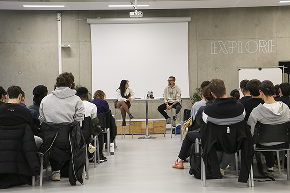 Un homme et une femme sont assis à une table haute en fond et devant eux il y a des jeunes qui les écoutent et qui sont assis.