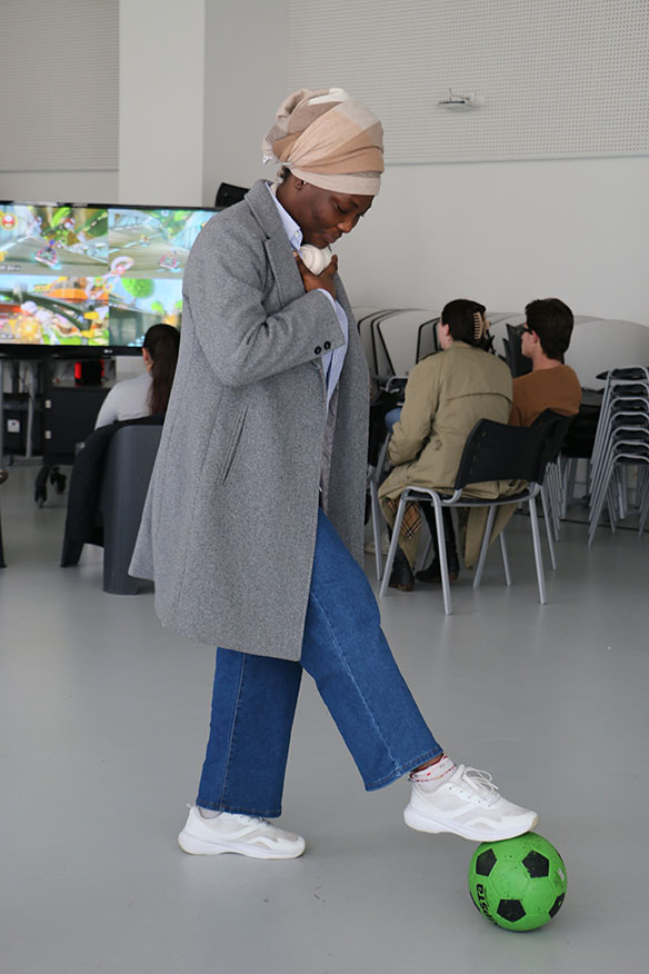 Etudiante qui a un ballon sous le pieds et un écran en fond sur lequel d'autres étudiants jouent à Mario kart.