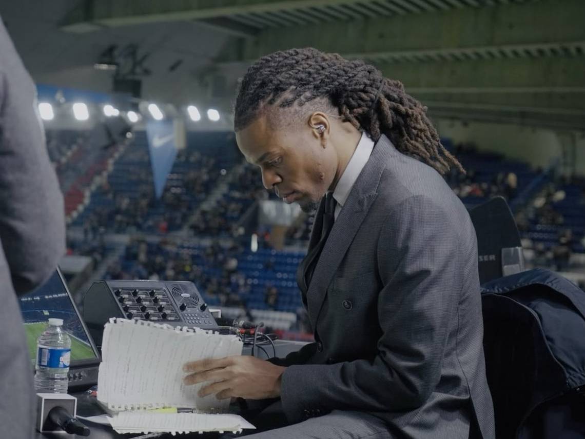 Loïc Rémy en costume relit ses notes en cabine de commentateur dans un stade